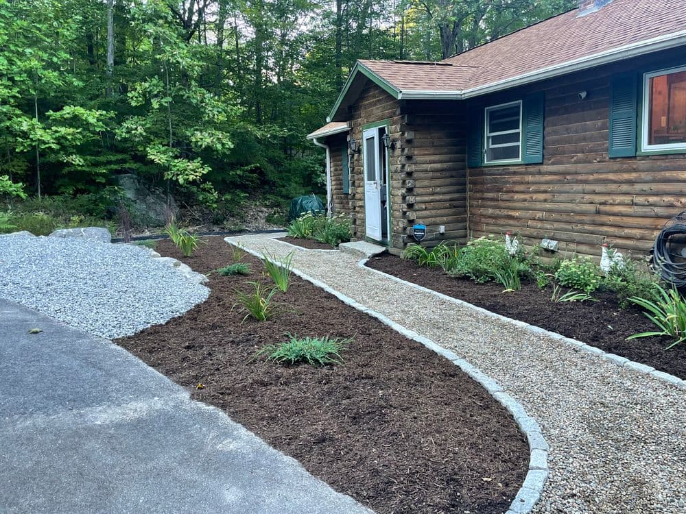 Log cabin home with landscaped garden, gravel path, and greenery in wooded area.