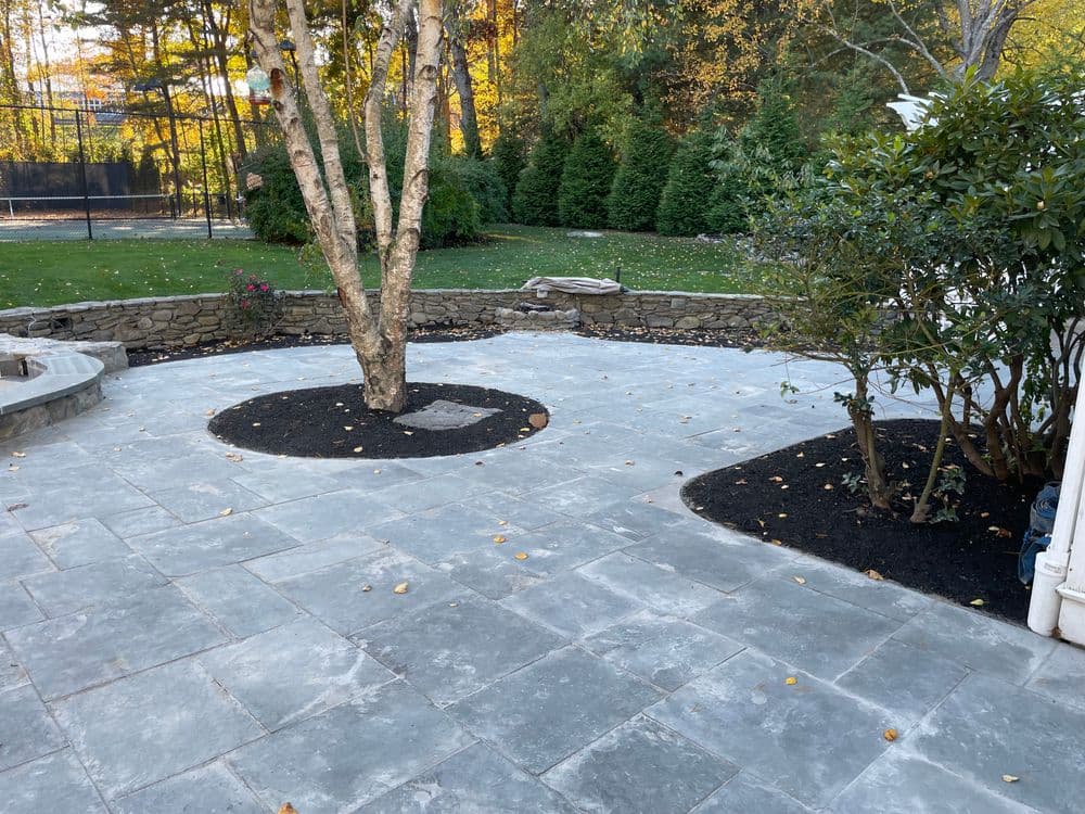 Patio with stone surface, surrounding trees, and mulched garden beds in a landscaped yard.