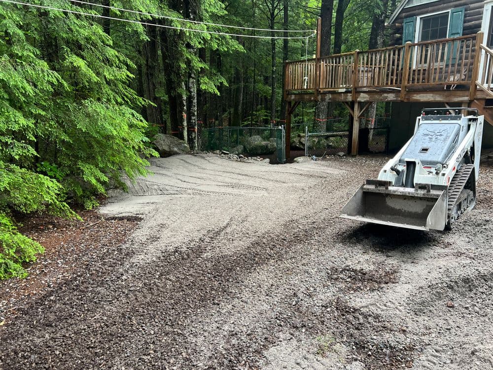 Excavator on gravel driveway with wooden deck in a forested area.