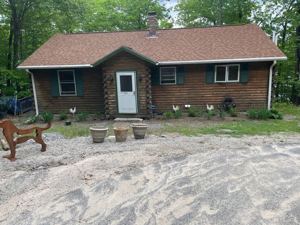Log cabin house with green shutters, gravel driveway, and flower beds in a wooded area.