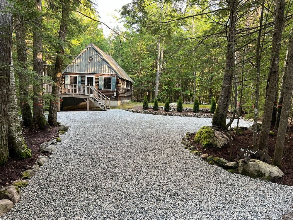 Charming log cabin nestled in a forest with a gravel driveway and landscaped garden.
