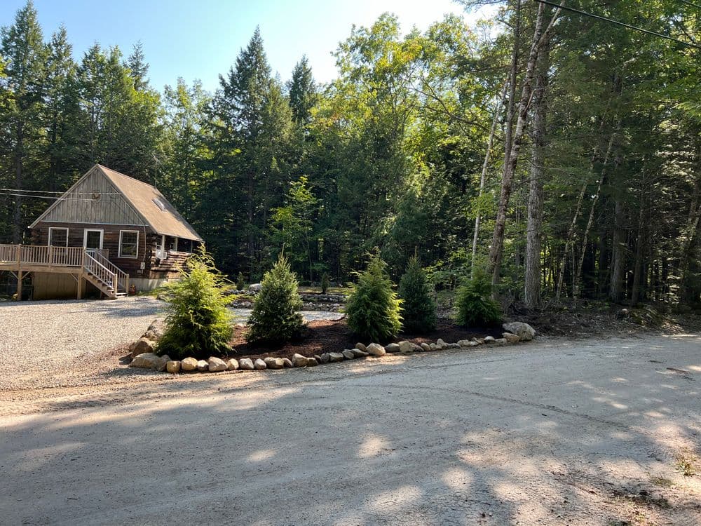 Cozy cabin surrounded by trees with landscaped yard and gravel driveway. Ideal for nature retreat.