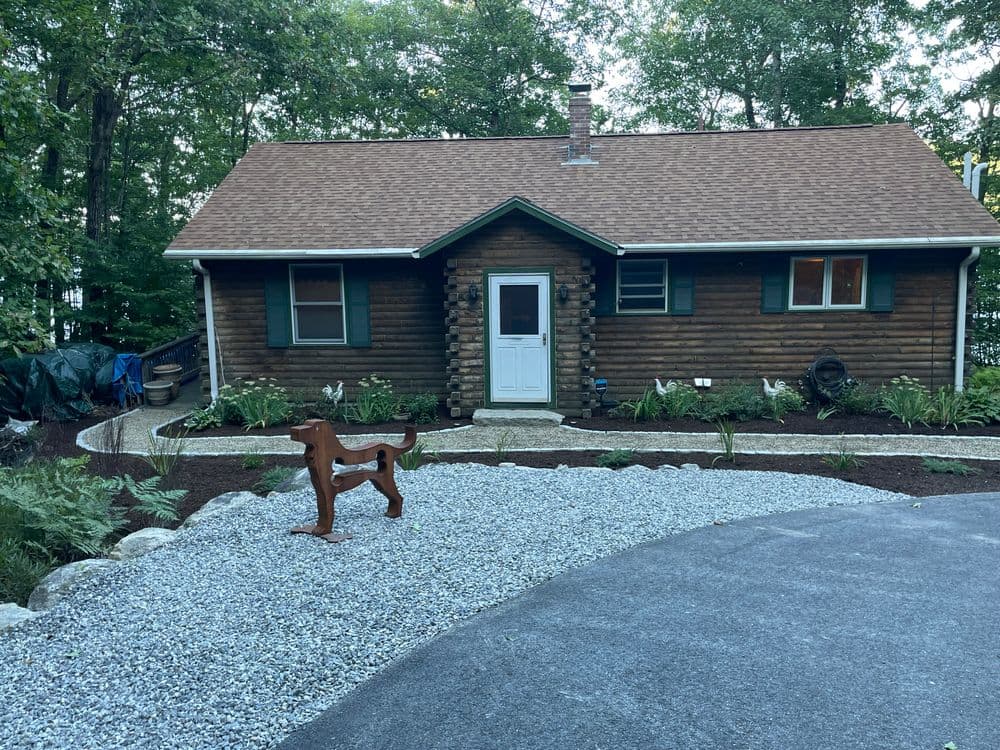Log cabin surrounded by greenery with a gravel path and wooden dog sculpture in front.