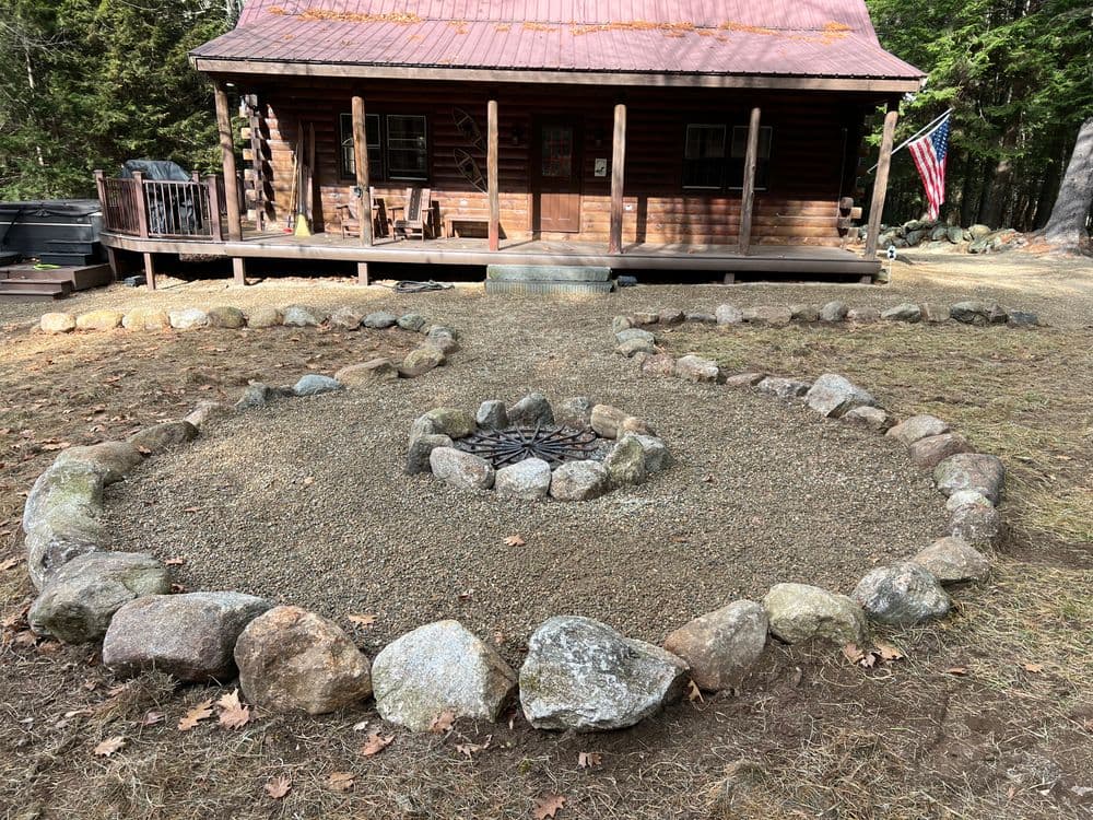 Log cabin with a stone fire pit surrounded by a circular rock arrangement and an American flag.