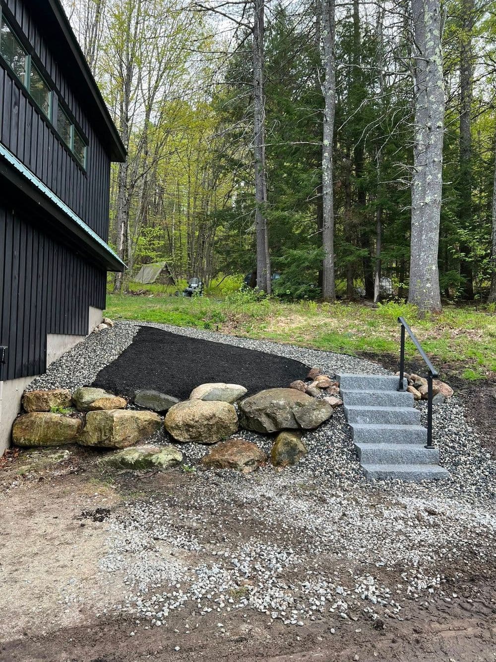 Gravel pathway with stone steps and landscaping by a black house in a wooded area.