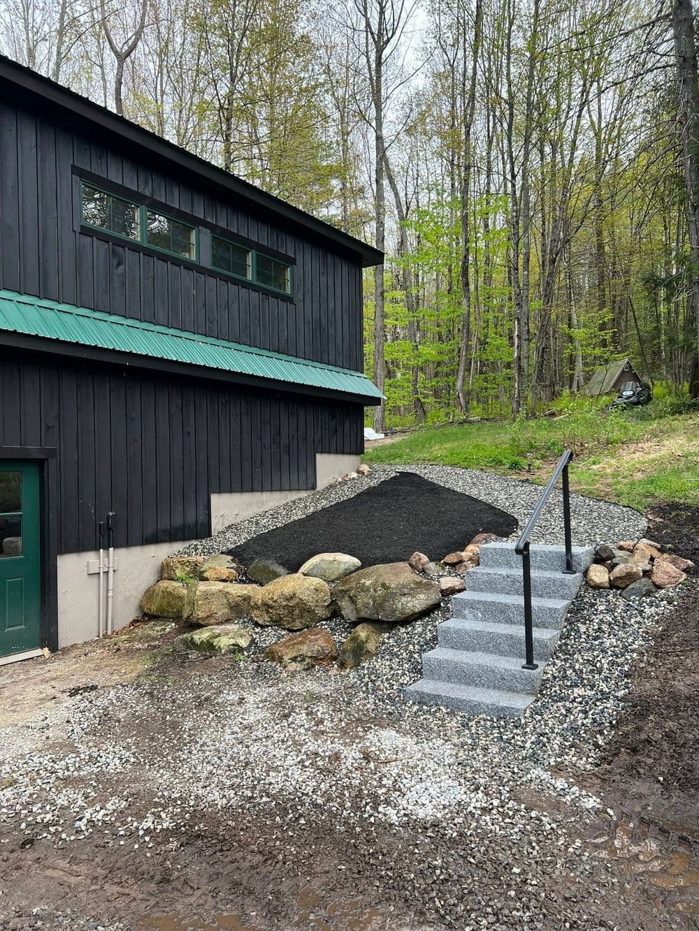 Modern black building with stairs, landscaped gravel, and natural stone features in wooded area.