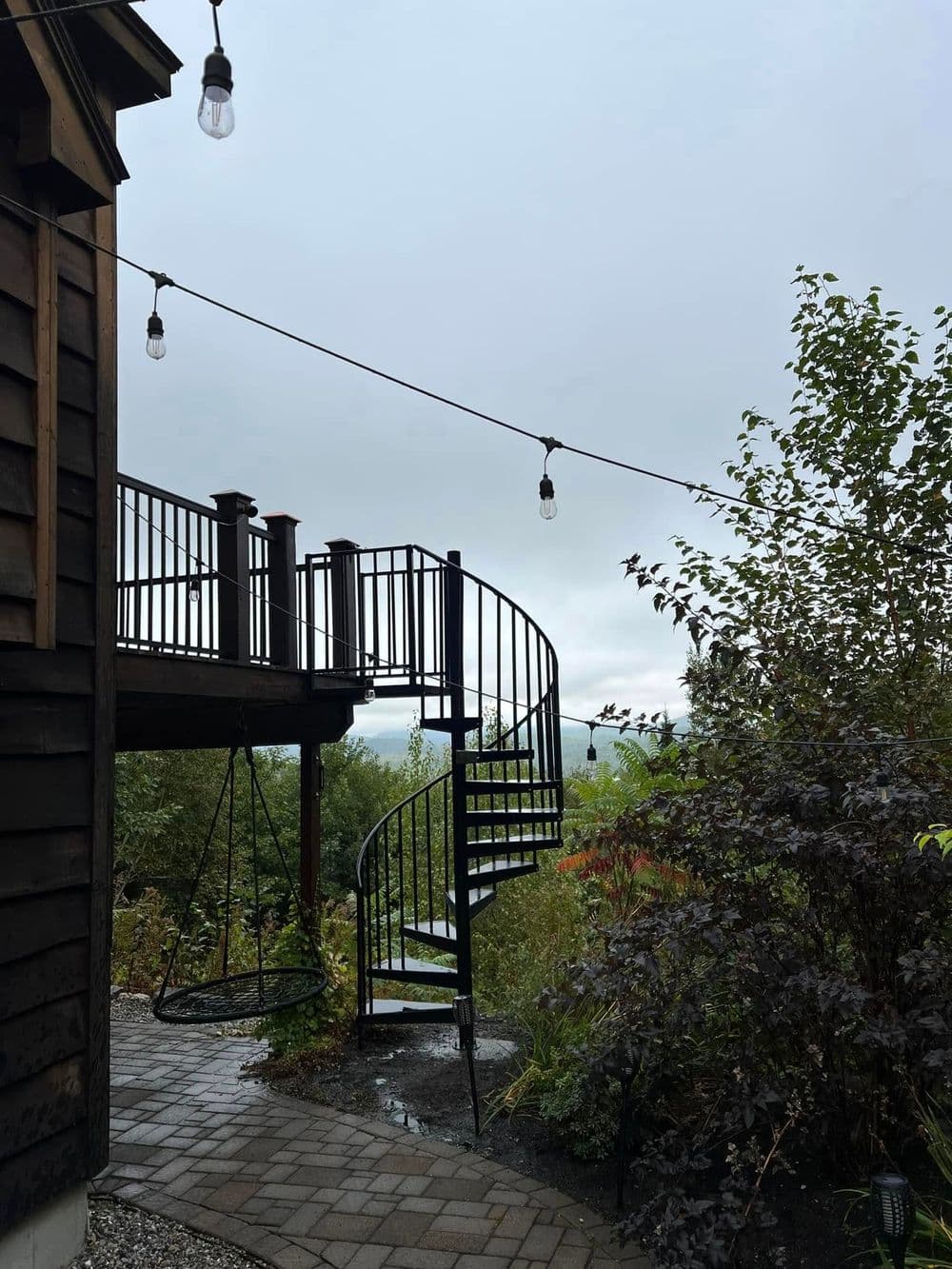 Spiral staircase leading to a deck surrounded by greenery on a cloudy day.