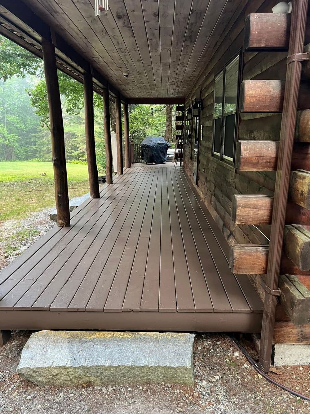 Brown wooden deck of a rustic cabin under a porch surrounded by greenery.
