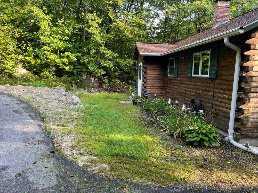 Cottage surrounded by greenery, featuring a gravel driveway and flower beds.